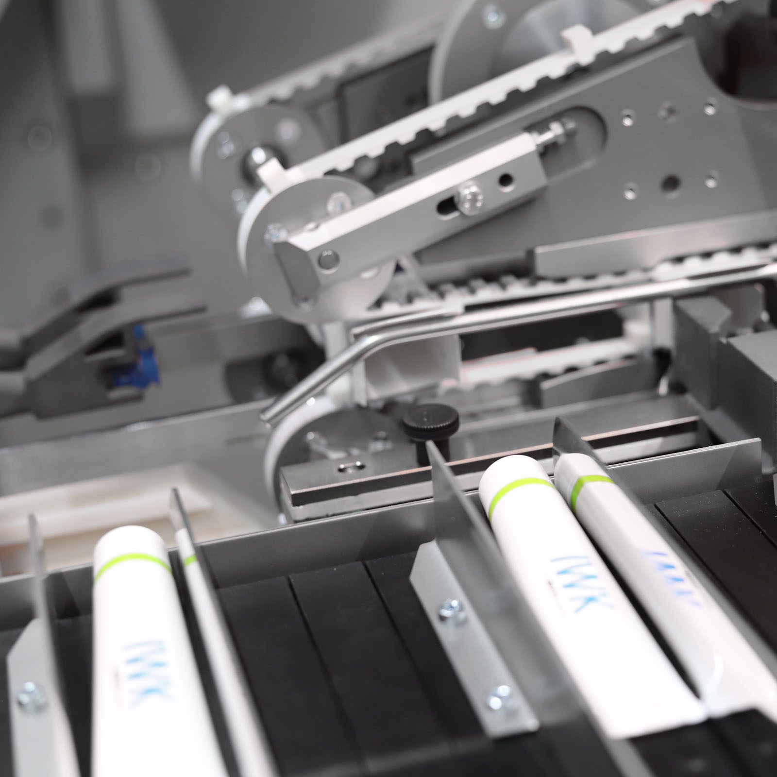 Tuben mit IWK‑Kennzeichnung in einer präzisen mechanischen Transport- und Zuführstrecke einer Produktionsanlage.