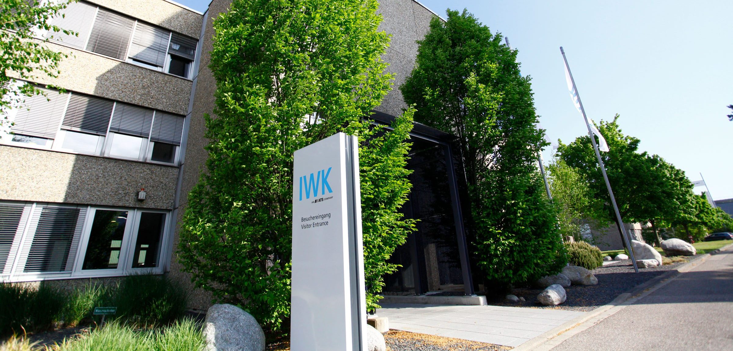 Eingangsbereich des IWK‑Firmengebäudes mit Firmenstele und umliegender Begrünung.