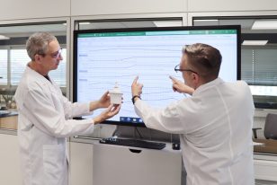 People in the laboratory analyze packaging components and discuss measurement results on a large monitor.