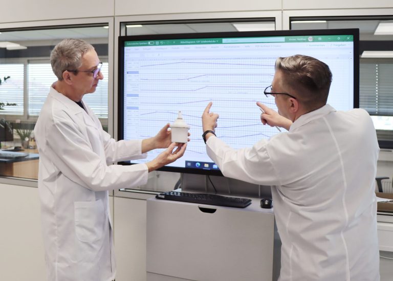 Personen im Labor analysieren Verpackungskomponenten und diskutieren Messergebnisse auf einem großen Monitor.