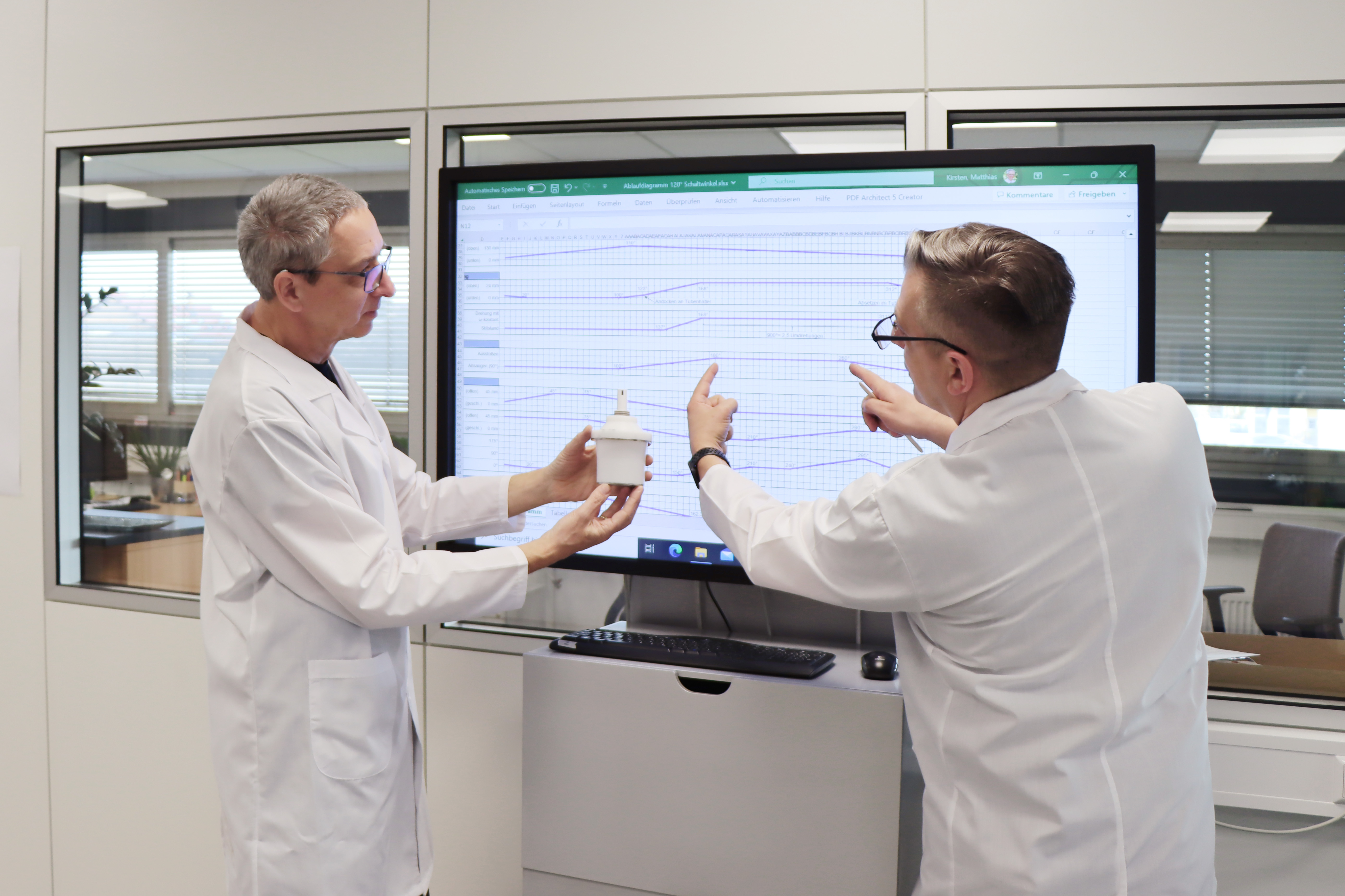 People in the laboratory analyze packaging components and discuss measurement results on a large monitor.