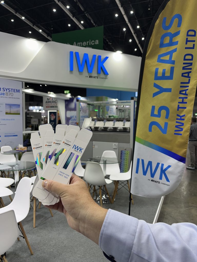 Hand holds several IWK giveaways in front of the IWK booth with anniversary flag "25 Years IWK Thailand Ltd".